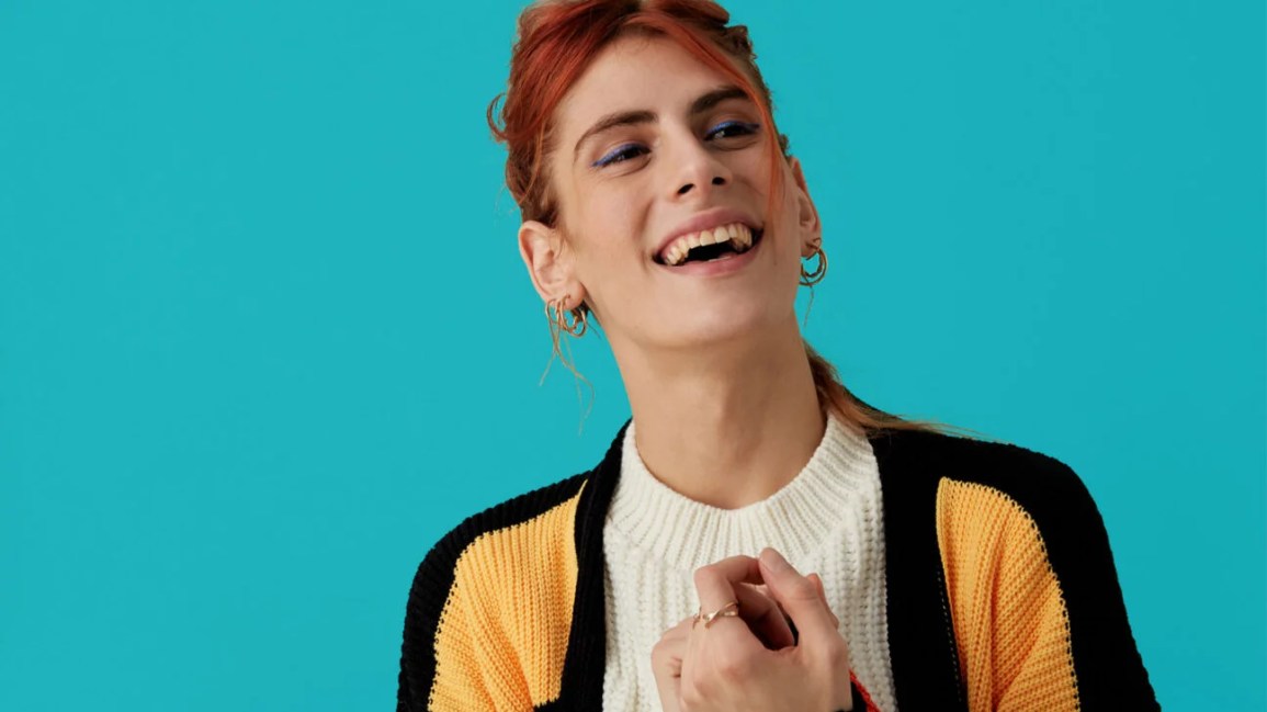 cropped image of a trans woman wearing a white blouse and black and yellow color-blocked cardigan smiling at something off screen