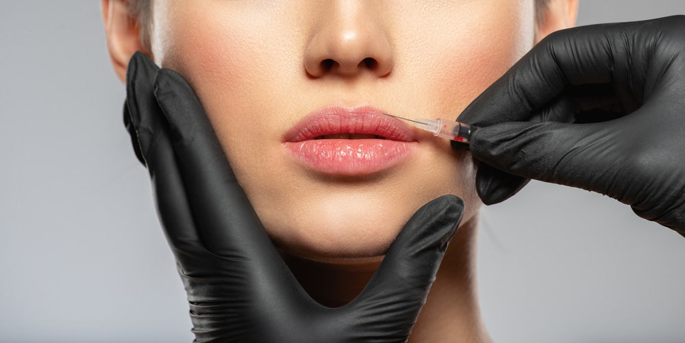 A woman has an injection administered to her lips by a black gloved hand as the other gloved hand holds her chin stable